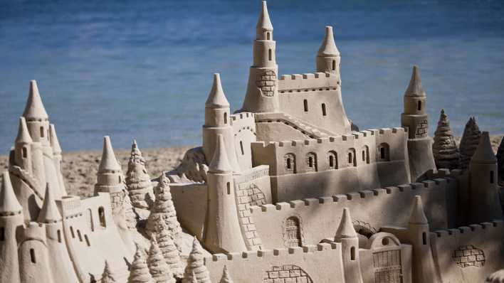 Eine Sandburg mit mehreren Türmen, im Hintergrund das blaue Meer (Quelle: imago/JuNiArt)